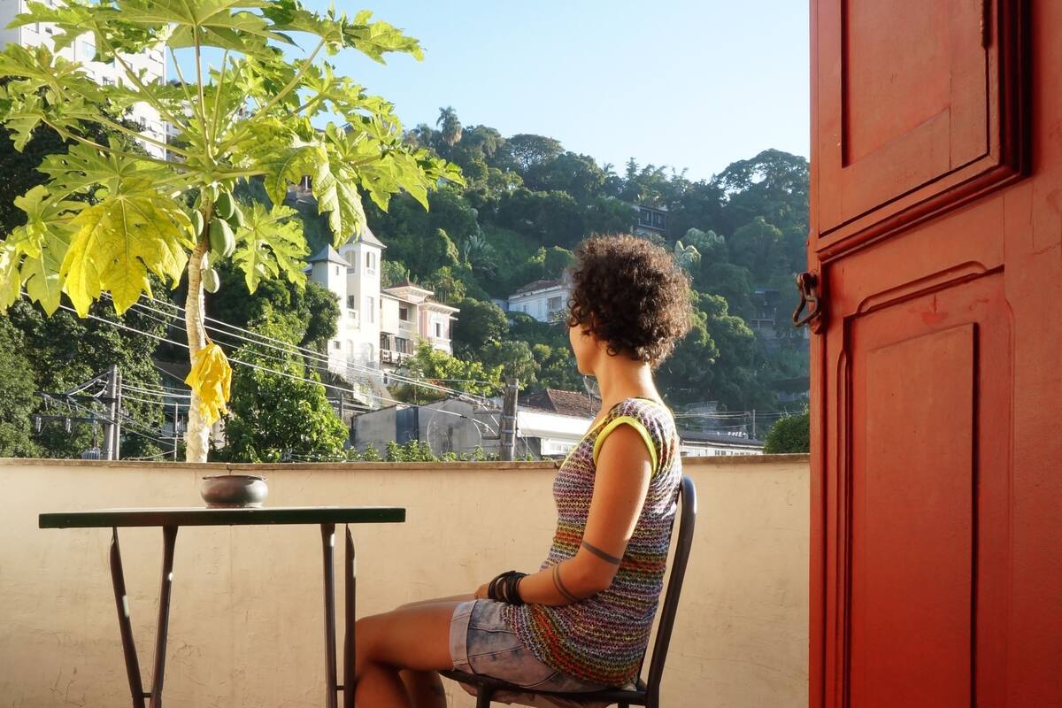 Eine Person sitzt an einem Tisch auf einem Balkon mit Blick auf Gebäude und Bäume.