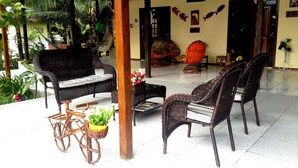 Un espacio interior amplio con mobiliario de mimbre, incluyendo un sofá y sillas, una mesita y una butaca decorativa. Se observan plantas en macetas y una hamaca al fondo.