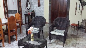 Una sala de estar con dos sillones de mimbre, una mesa de centro de vidrio con un florero, una mesa de comedor de madera con sillas y un piso de baldosas.