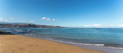 A beach with clear blue water, sandy shore, and distant hills.