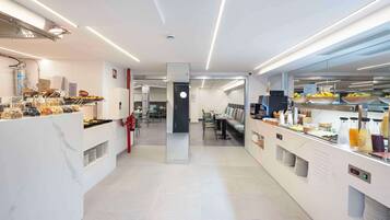 Un área de desayuno moderna y bien iluminada, con diversas opciones de comida y bebida, un espacio de estar y una cocina visible al fondo.
