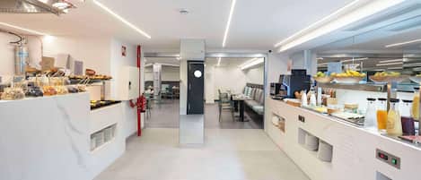 A modern, well-lit breakfast area with a variety of food and drink options, a seating area, and a visible kitchen in the background.