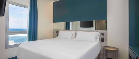 A modern hotel room with a large bed, a small table, a TV, and a view of the ocean.