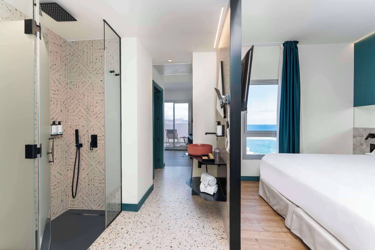 Habitación de hotel con cama, baño con ducha, lavabo y ventana con vista al mar.