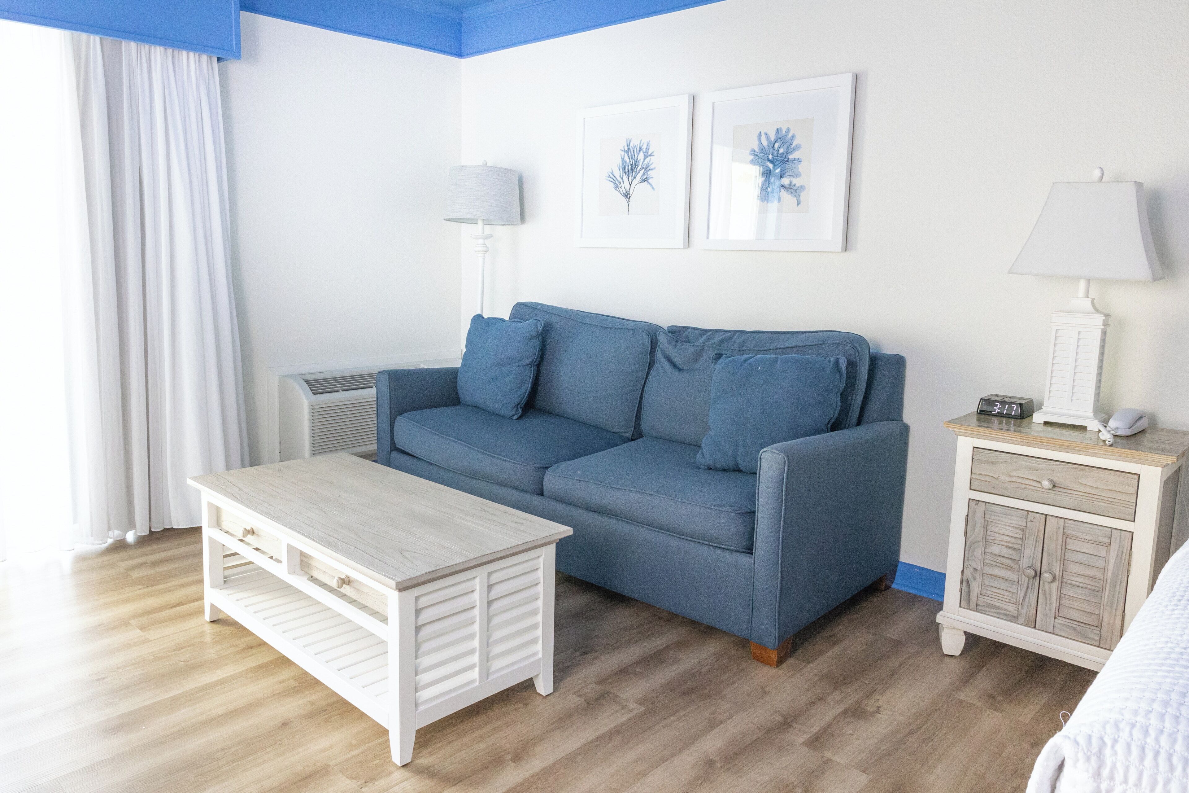 Una habitación con un sofá azul, una mesa blanca, una mesita de noche con una lámpara y un aire acondicionado instalado en la pared.