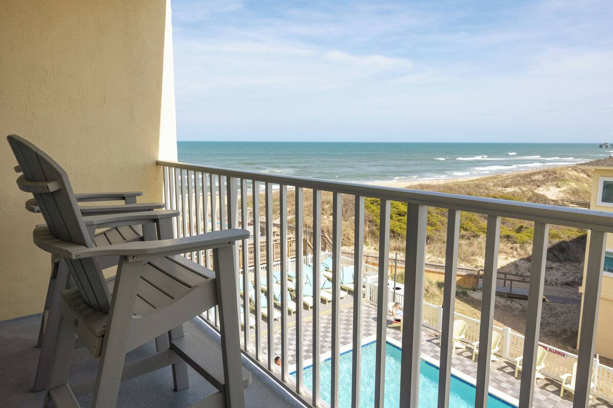 A balcony with a view of the ocean, a pool, and beach chairs.