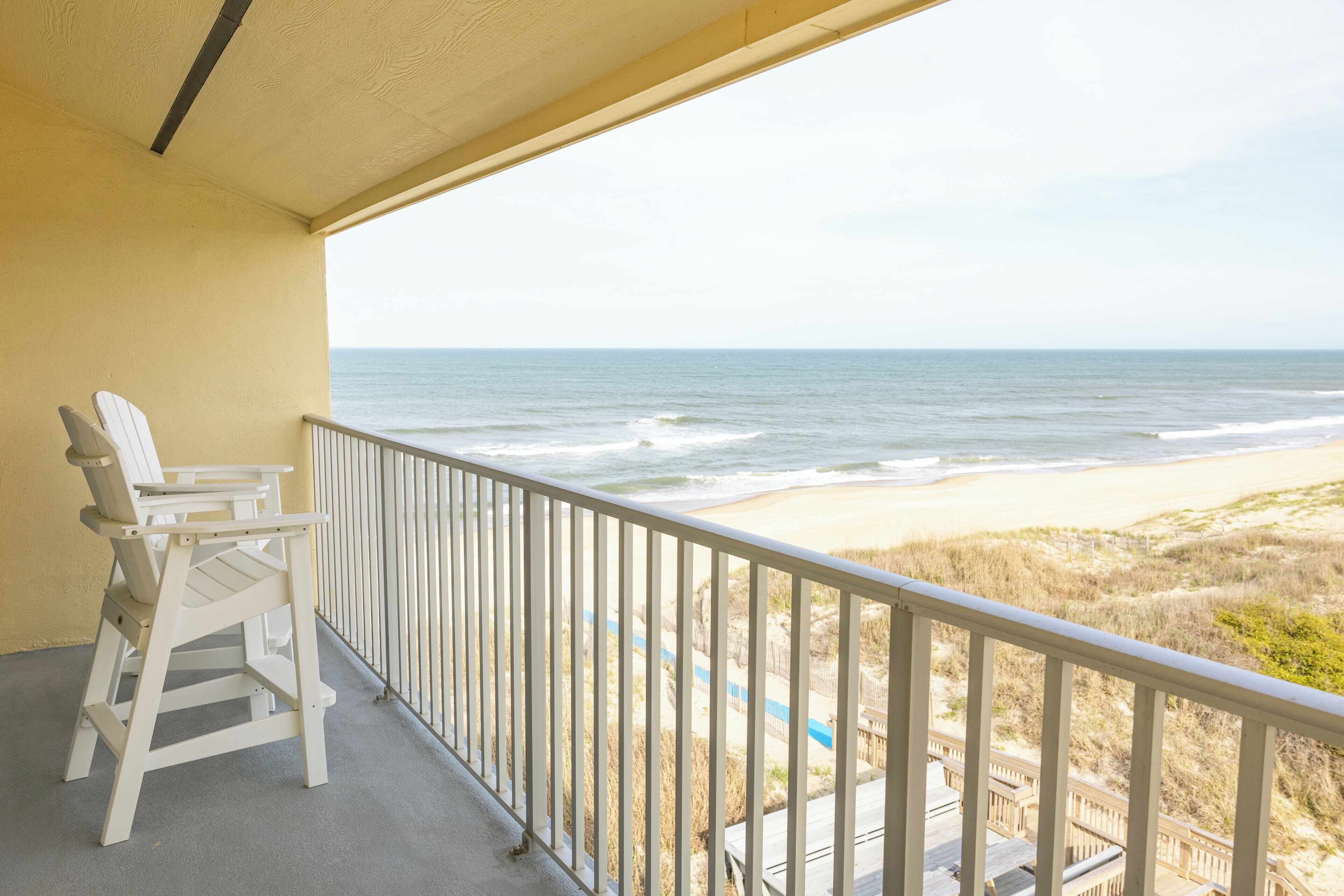 A balcony with a view of the ocean and beach, featuring two white chairs and a railing.