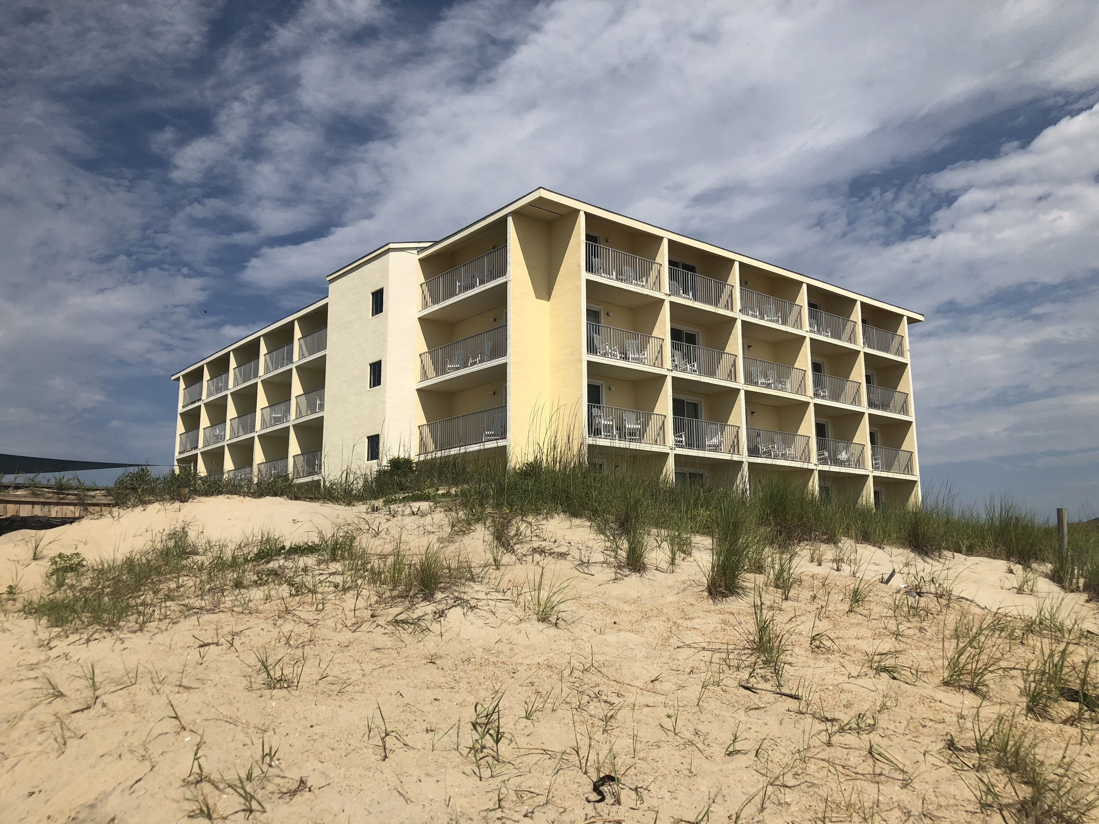 Un hotel de varios pisos con balcones, ubicado sobre una duna arenosa con vegetación escasa.
