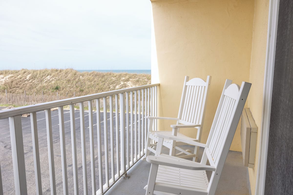 A balcony with two white chairs, a railing, and a view of the beach and dunes.