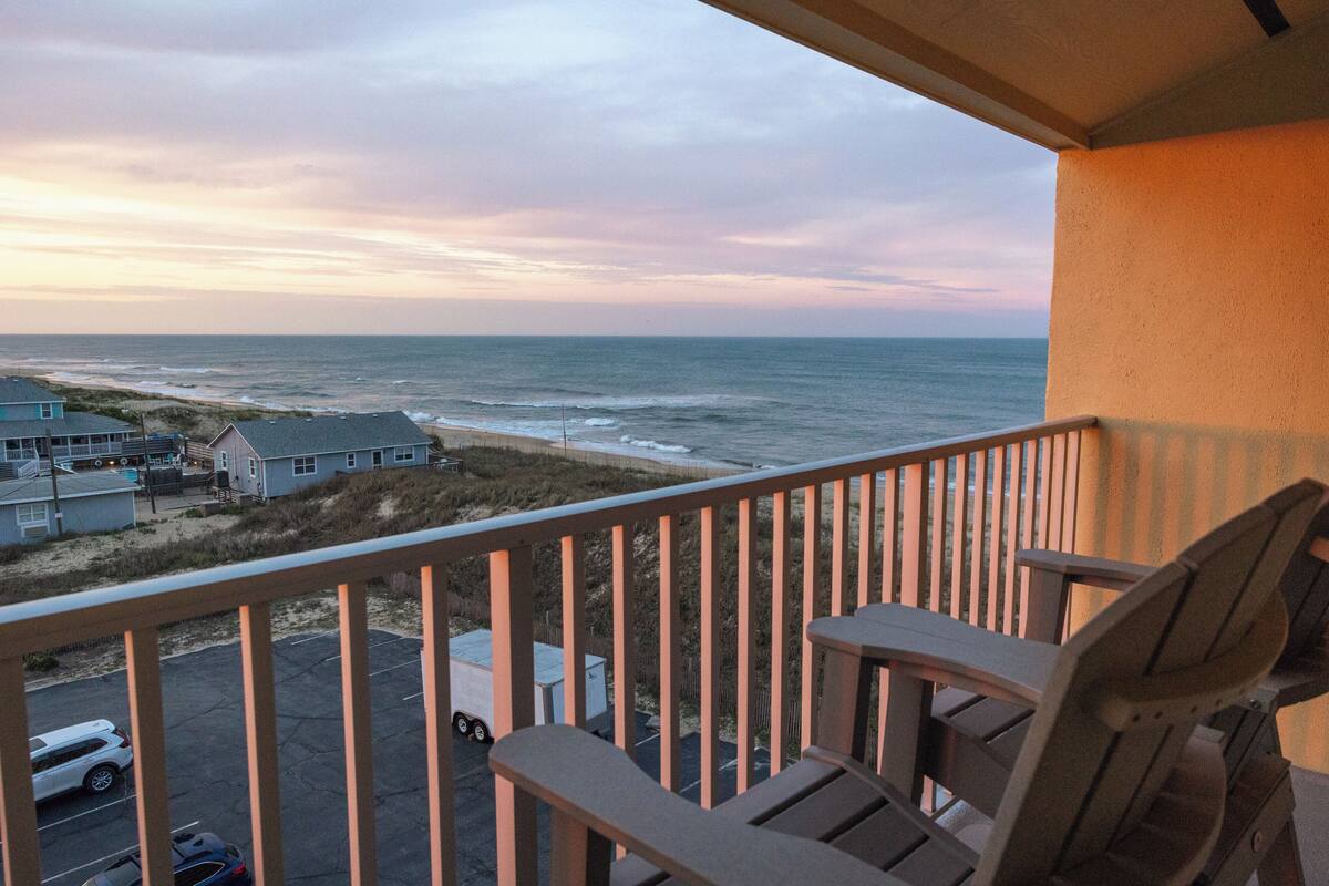 A balcony with a view of the ocean and beach, wooden chairs, and a parking area with a truck.