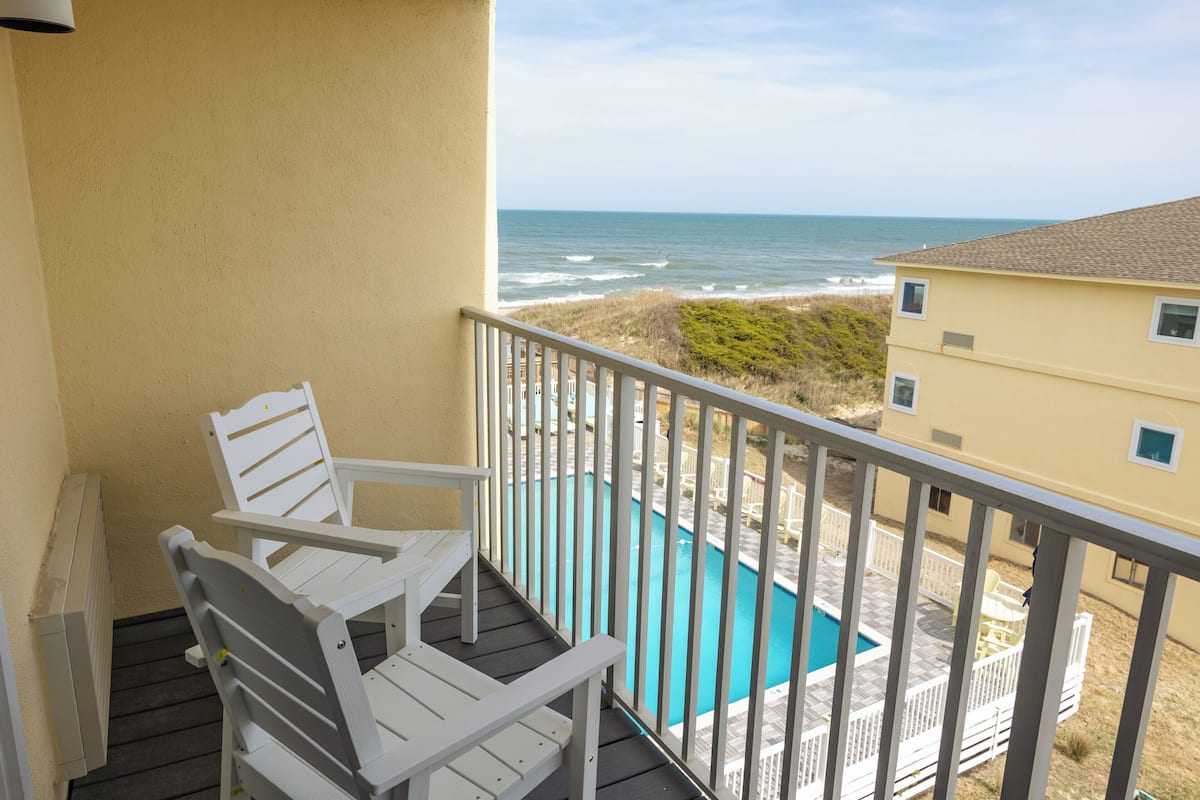 A balcony with white chairs, a view of the ocean, and a swimming pool.