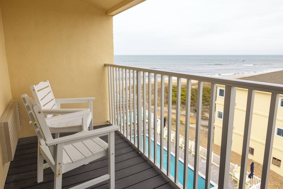 A balcony with a white chair, a pool, and a view of the beach.