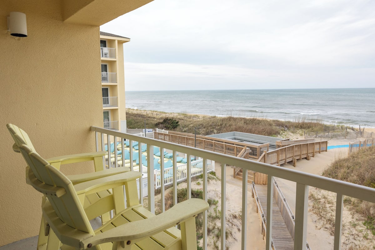A balcony with a view of the beach, a pool area, and a wooden deck.
