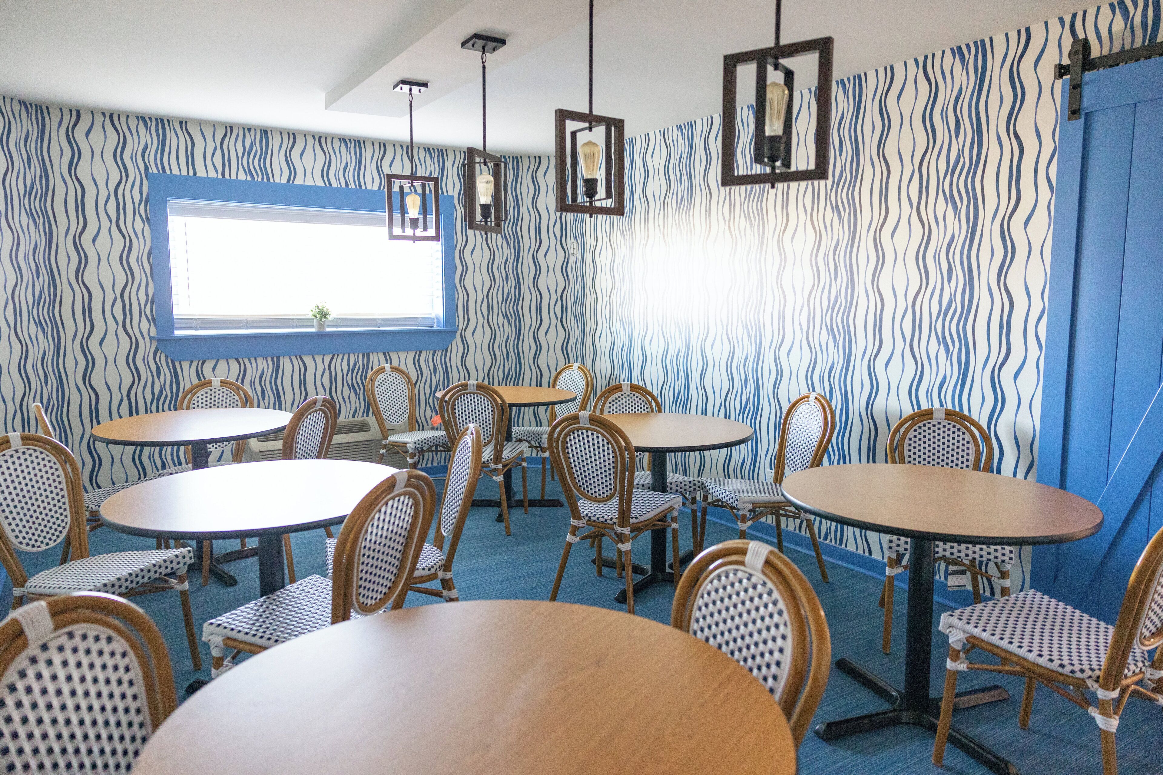 A room with round tables and wicker chairs, a window with blinds, and patterned wallpaper.