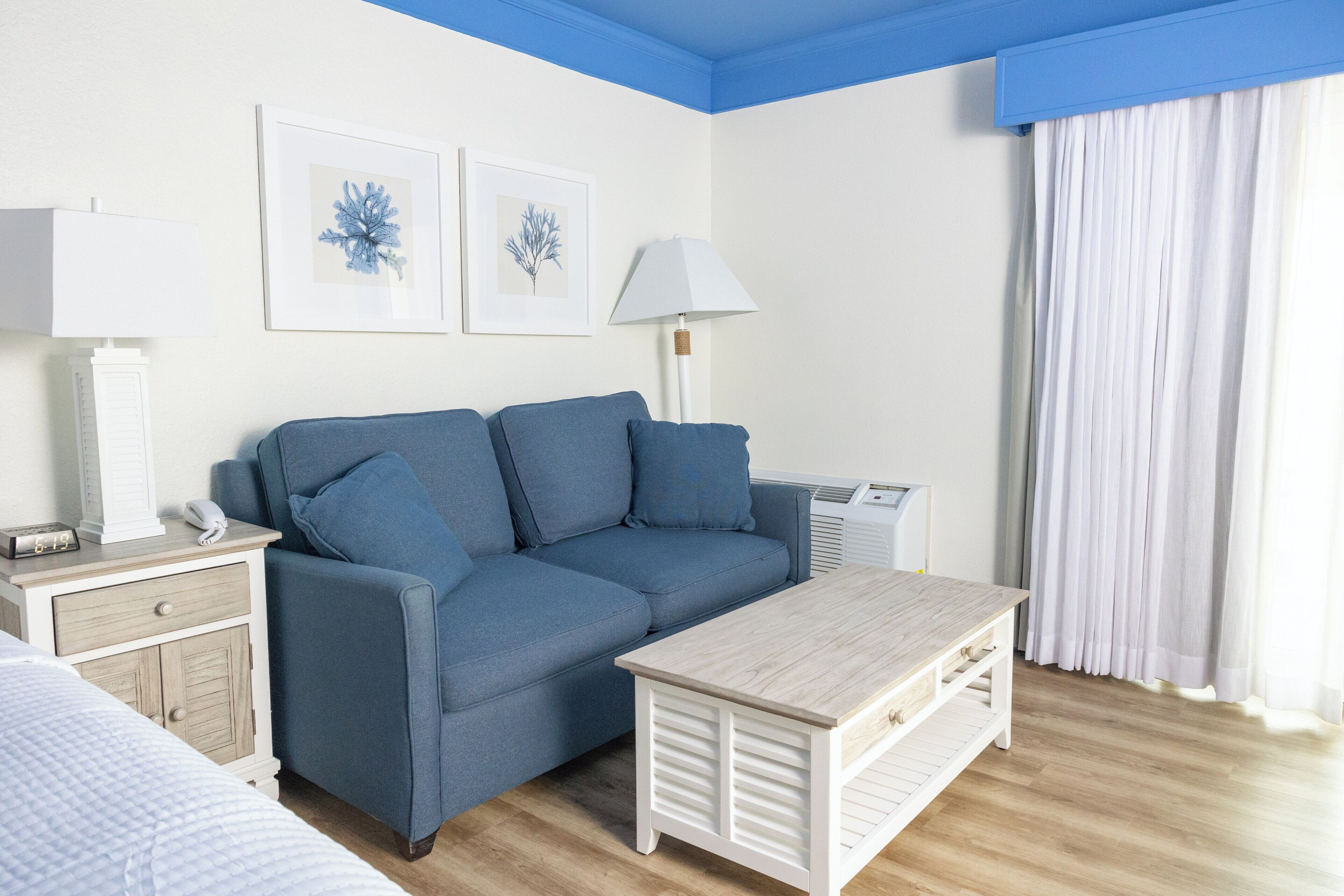 Una habitación con un sofá azul, una mesita de noche blanca, una lámpara y un cuadro en la pared.