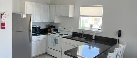 A kitchen with white cabinets, a stainless steel refrigerator, a stove, and a sink. There is a window with blinds and a telephone on the wall.