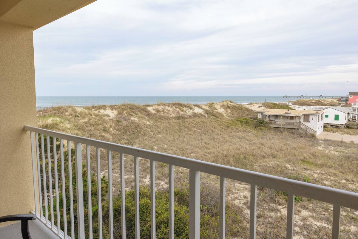 A balcony with a view of the beach, sandy dunes, and a pier in the distance.
