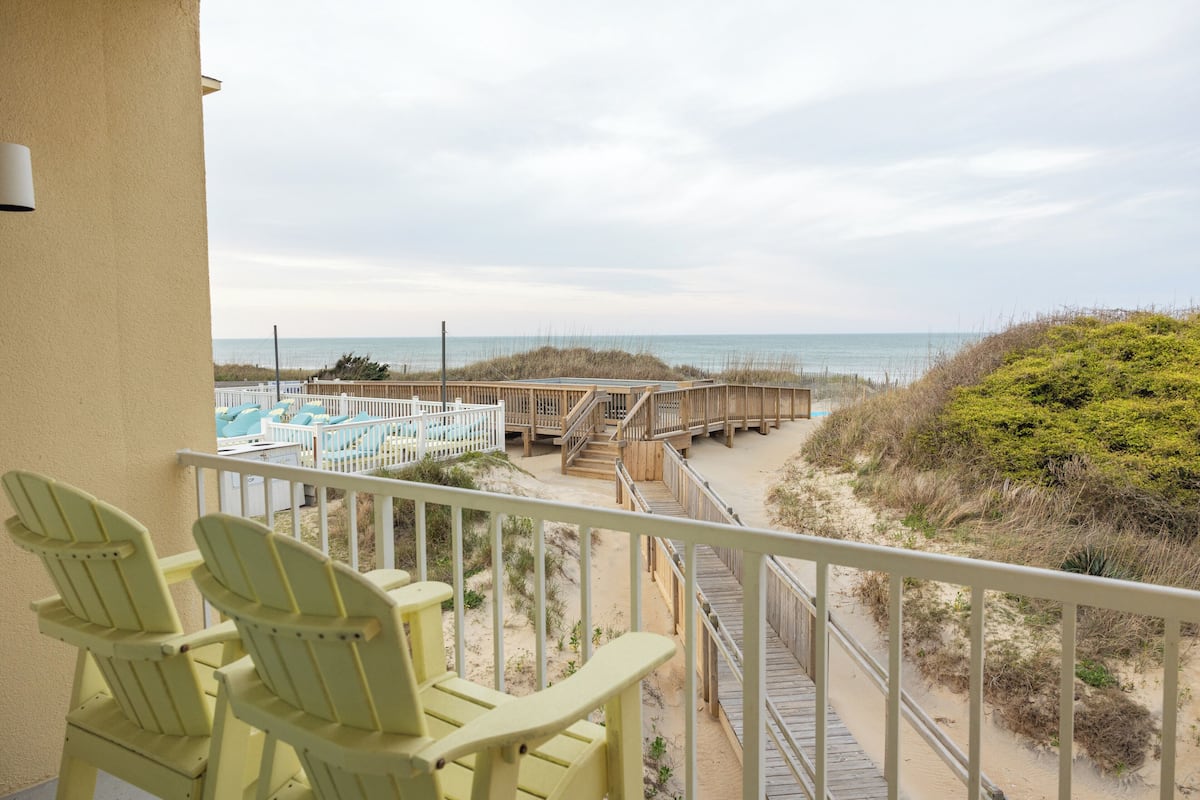 A balcony with yellow chairs, a wooden deck, and a view of the beach and ocean.