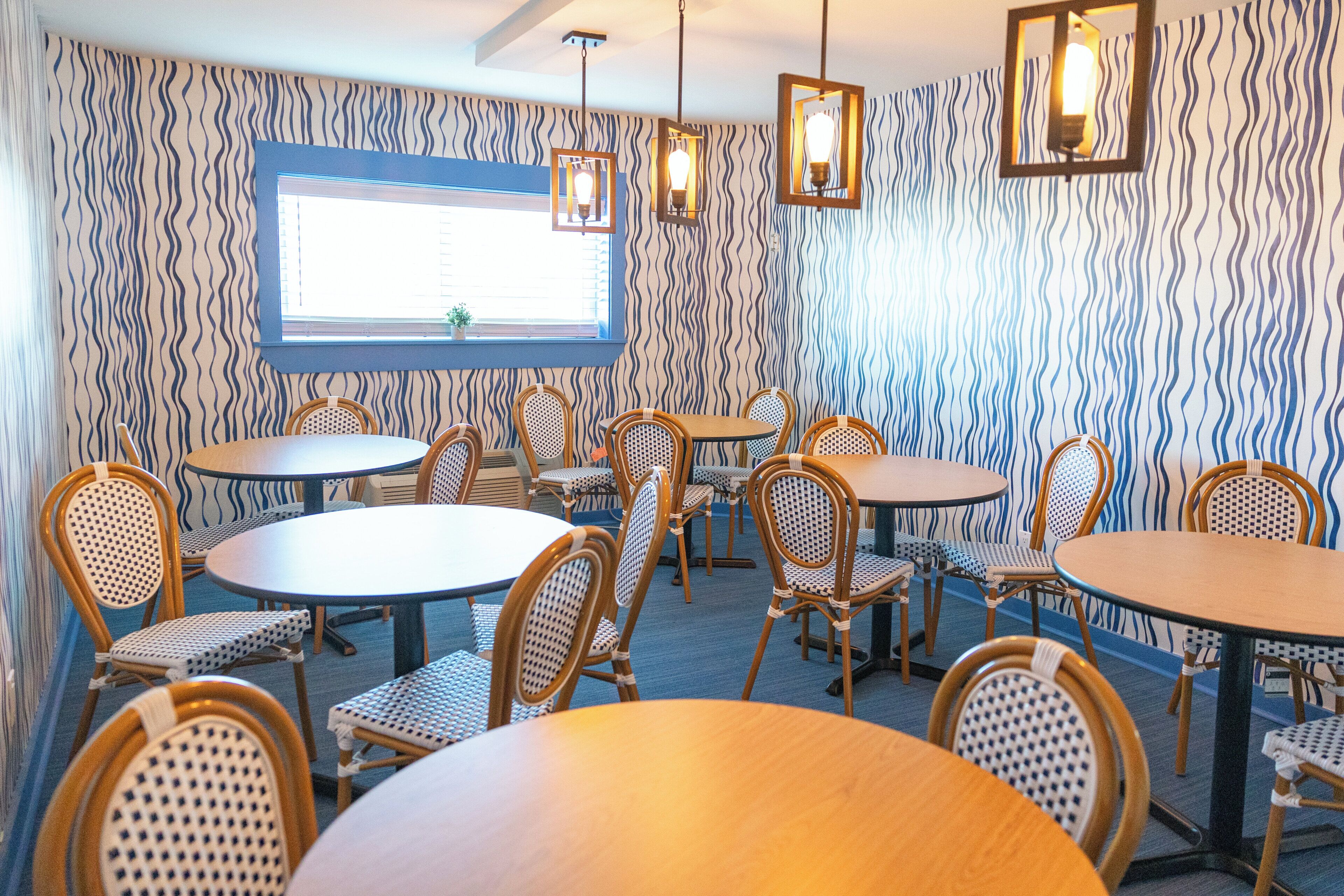 A dining area with round tables and wicker chairs, patterned wallpaper, and hanging pendant lights.