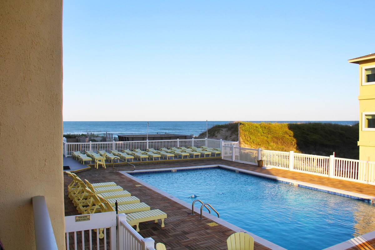 A swimming pool with a view of the ocean, surrounded by white railings and yellow lounge chairs.