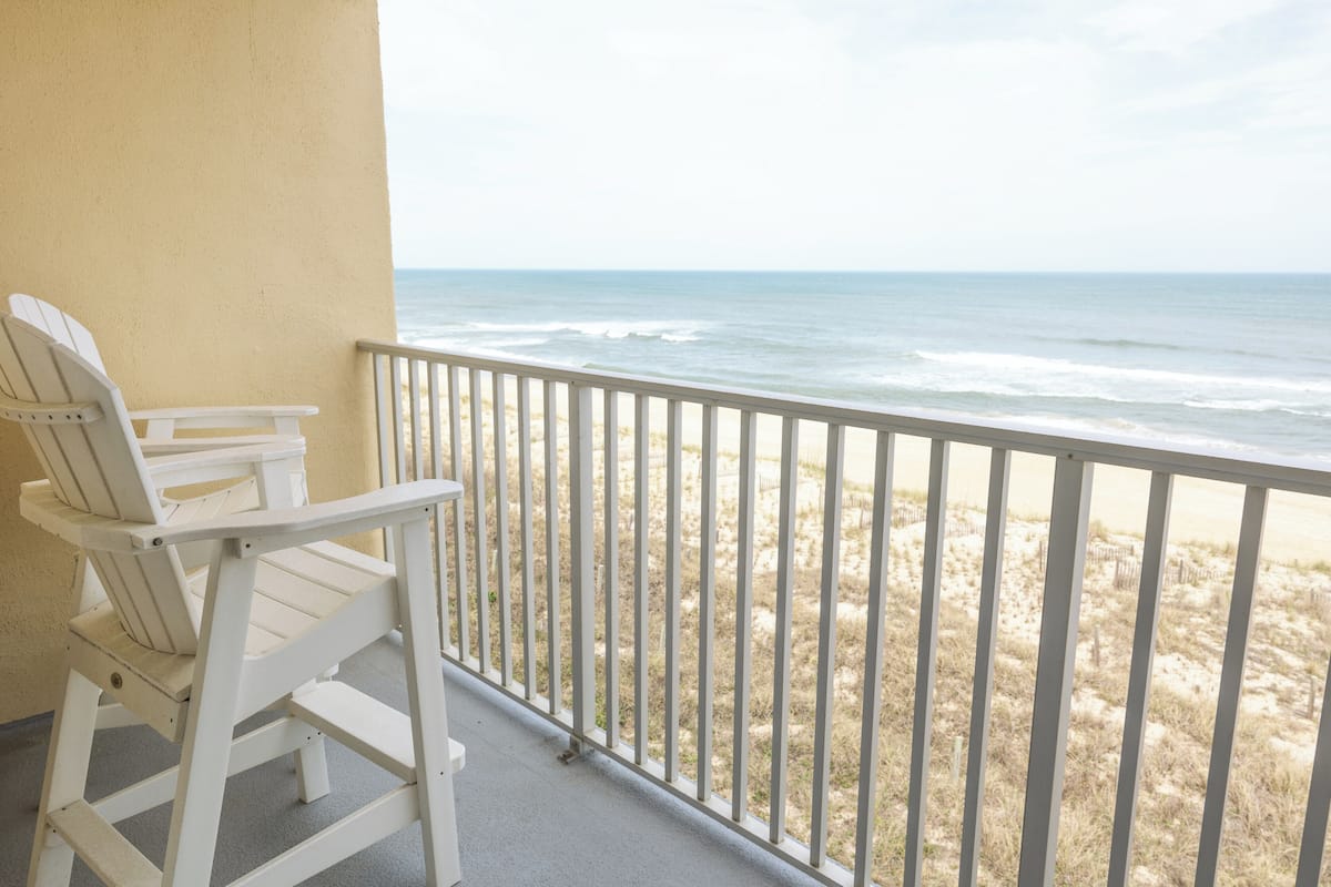 A balcony with a white chair, a view of the beach, and a railing.