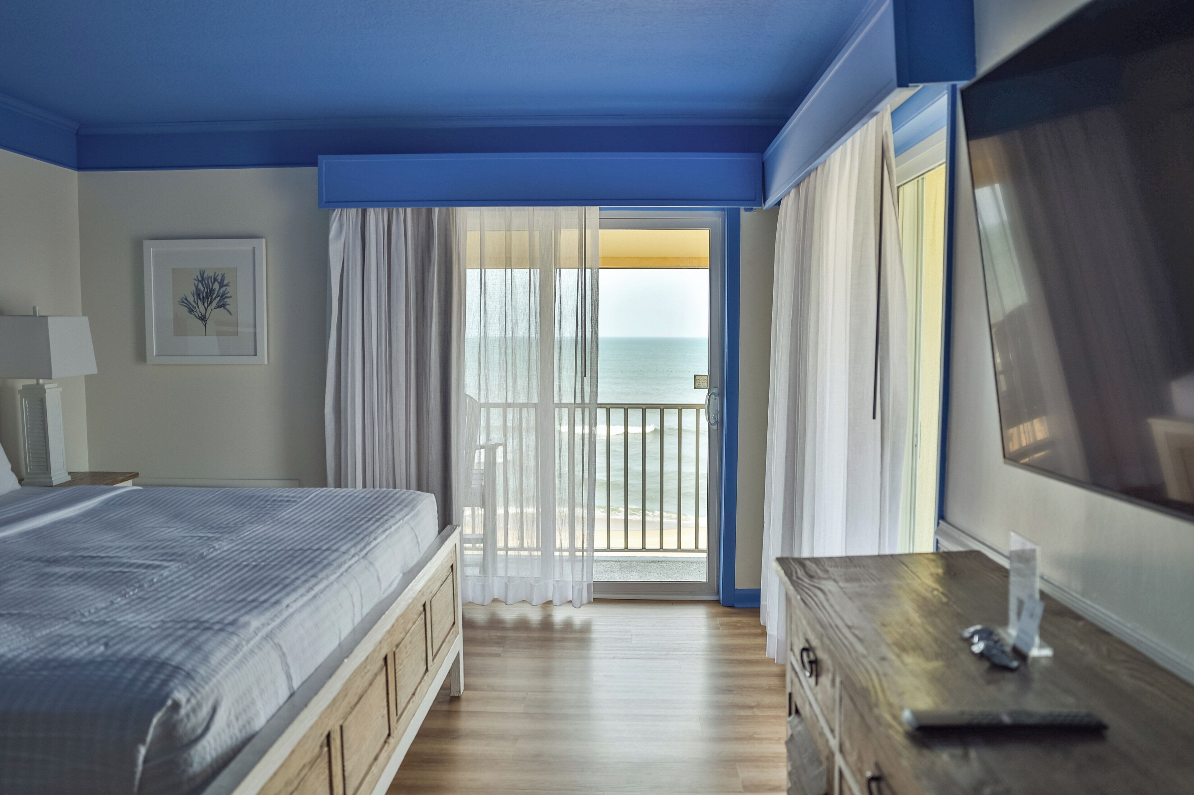 Une chambre à coucher comprenant un lit, une commode, un téléviseur, un tableau encadré et un balcon avec vue sur la mer.