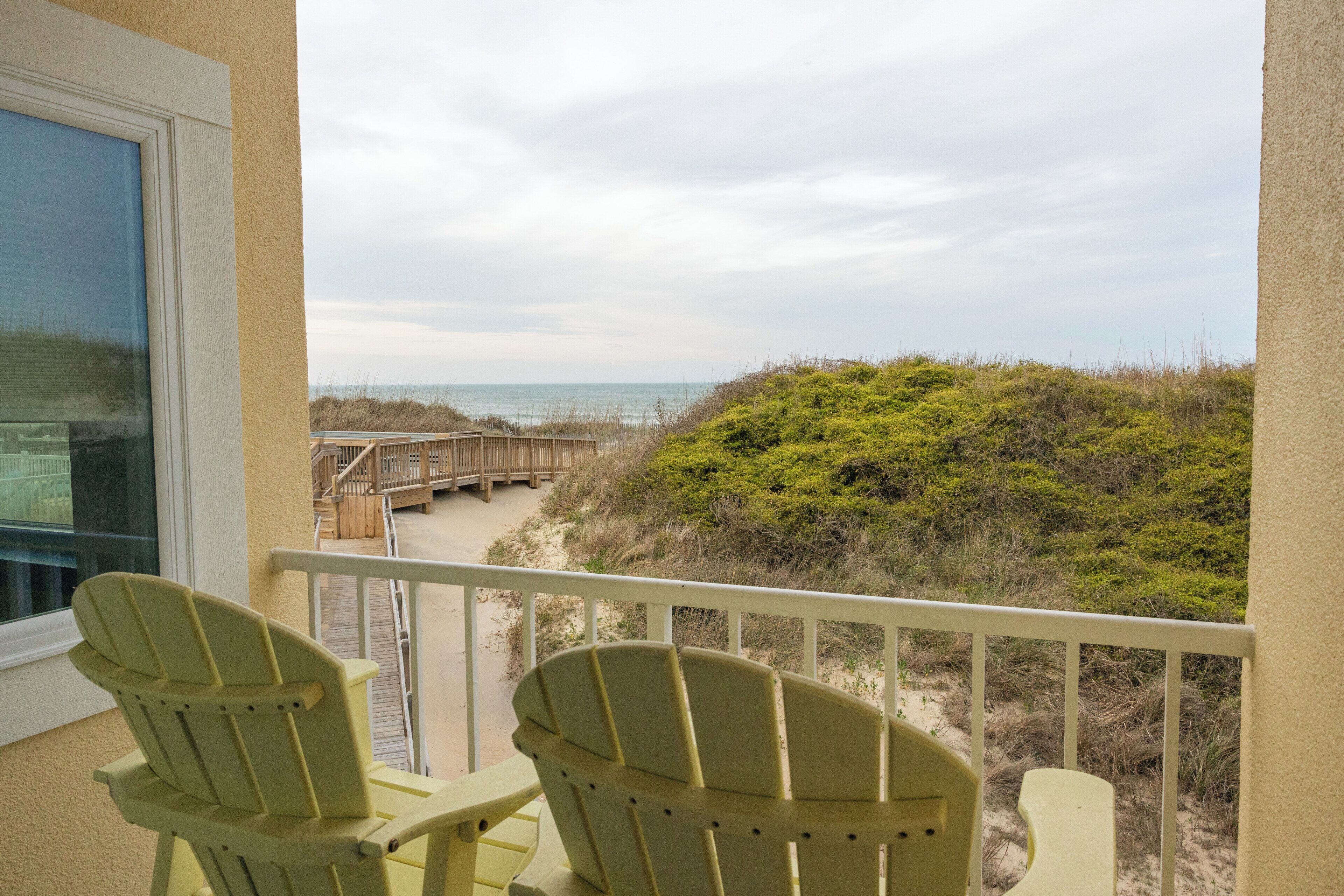 Un balcón con dos sillas, una terraza de madera y vistas a la playa y las dunas.
