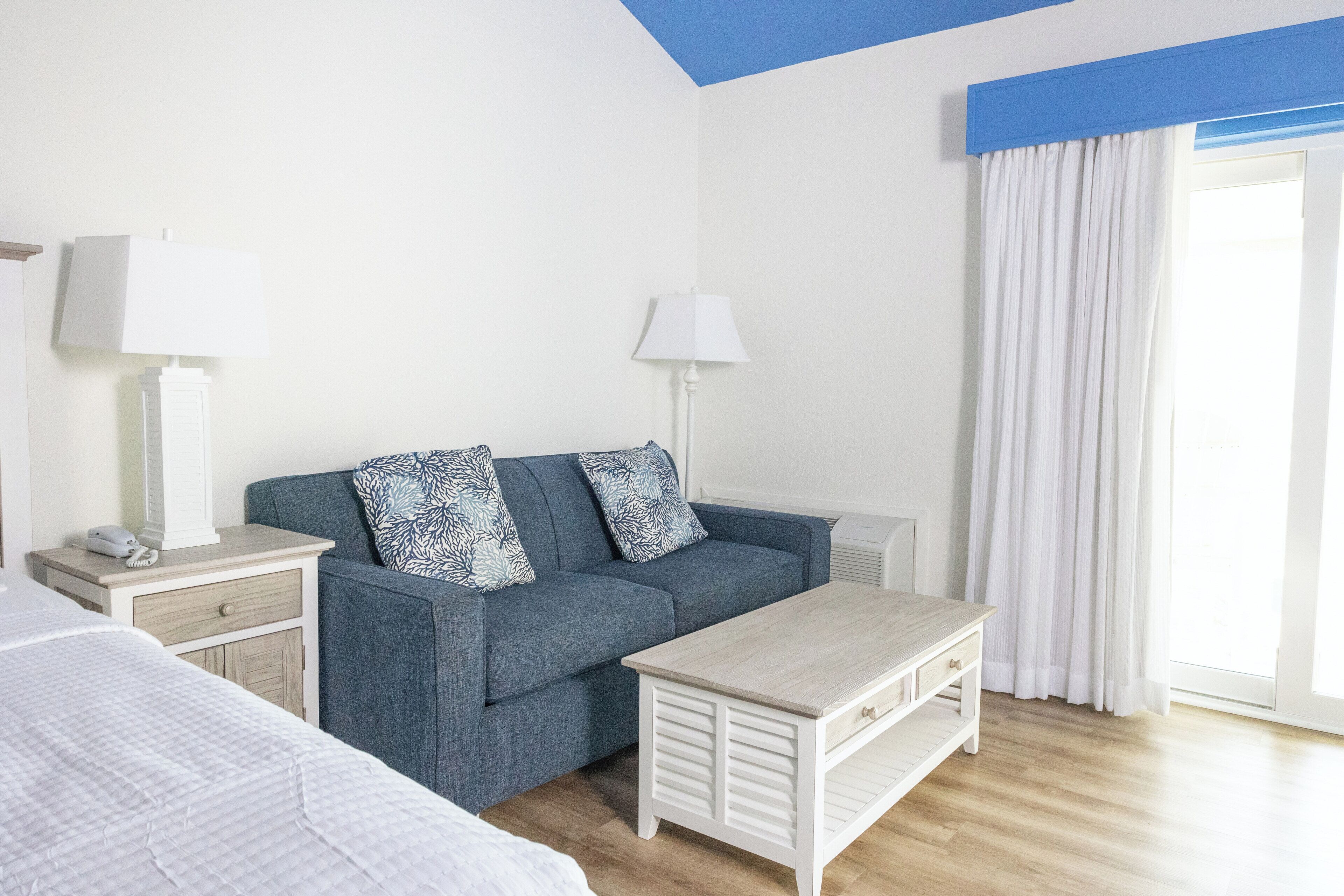 Et hotelværelse med en seng, en sofa, et lille bord, to sengeborde med lamper, et vindue med gardiner og et blåt listet loft.