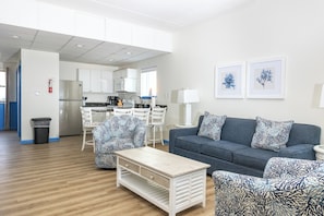 A modern living room with a grey sofa, patterned armchairs, a wooden coffee table, and a kitchen in the background.