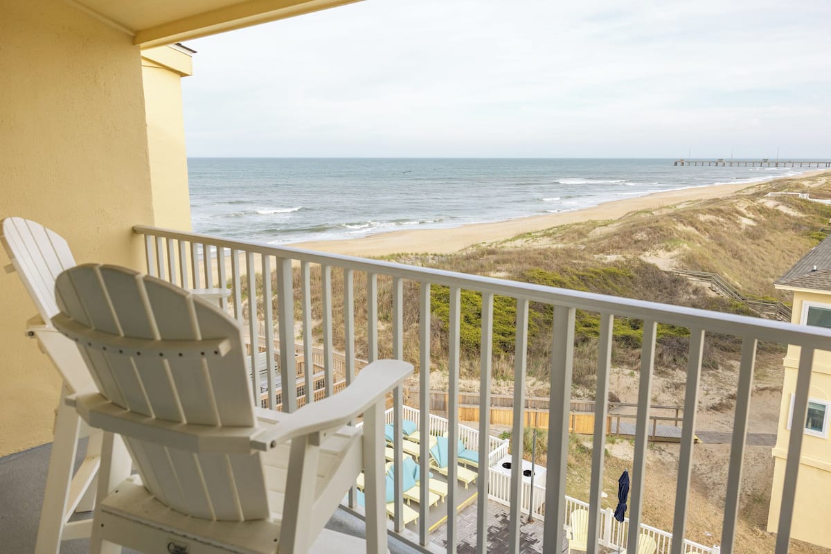 A balcony with a view of the beach and ocean, a white chair, and a wooden deck.