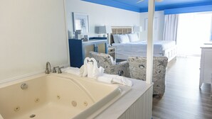 A hotel room with a large bathtub, a bed with white linens, a desk with a lamp, and a chair with a nautical pattern. - Surf Side Hotel (Nags Head)