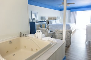 A hotel room with a large bathtub, a bed with white linens, a desk with a lamp, and a chair with a nautical pattern.