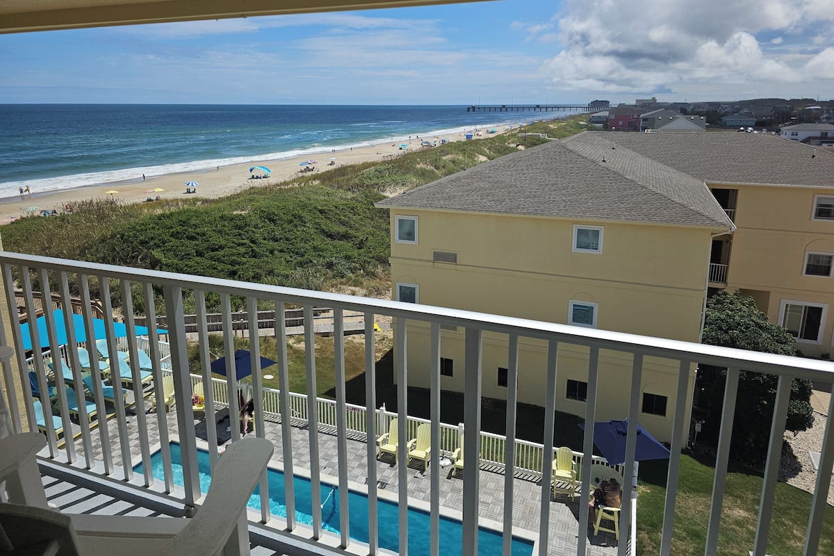 A balcony with a view of a beach, a pool, and a building with multiple windows.