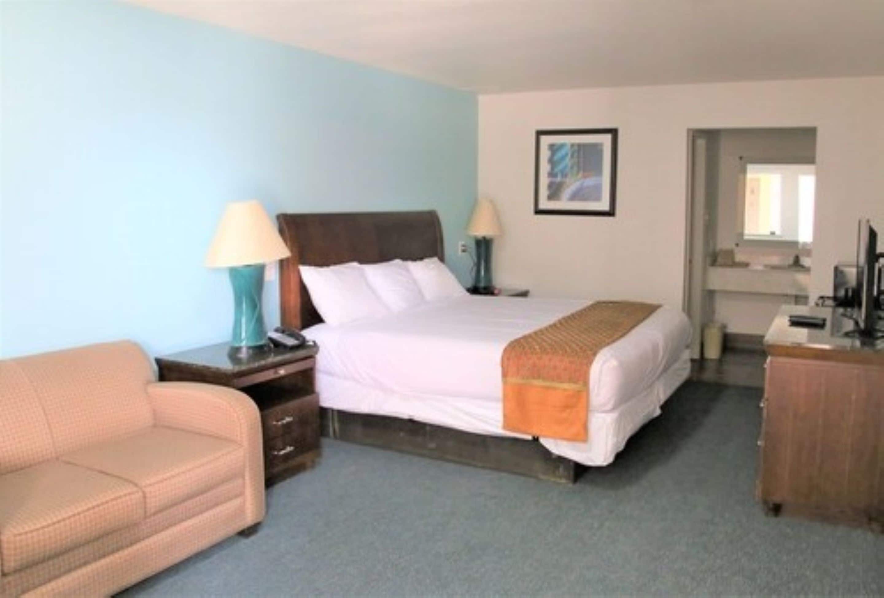 A hotel room with a bed, a nightstand, a lamp, a dresser, and a sofa.