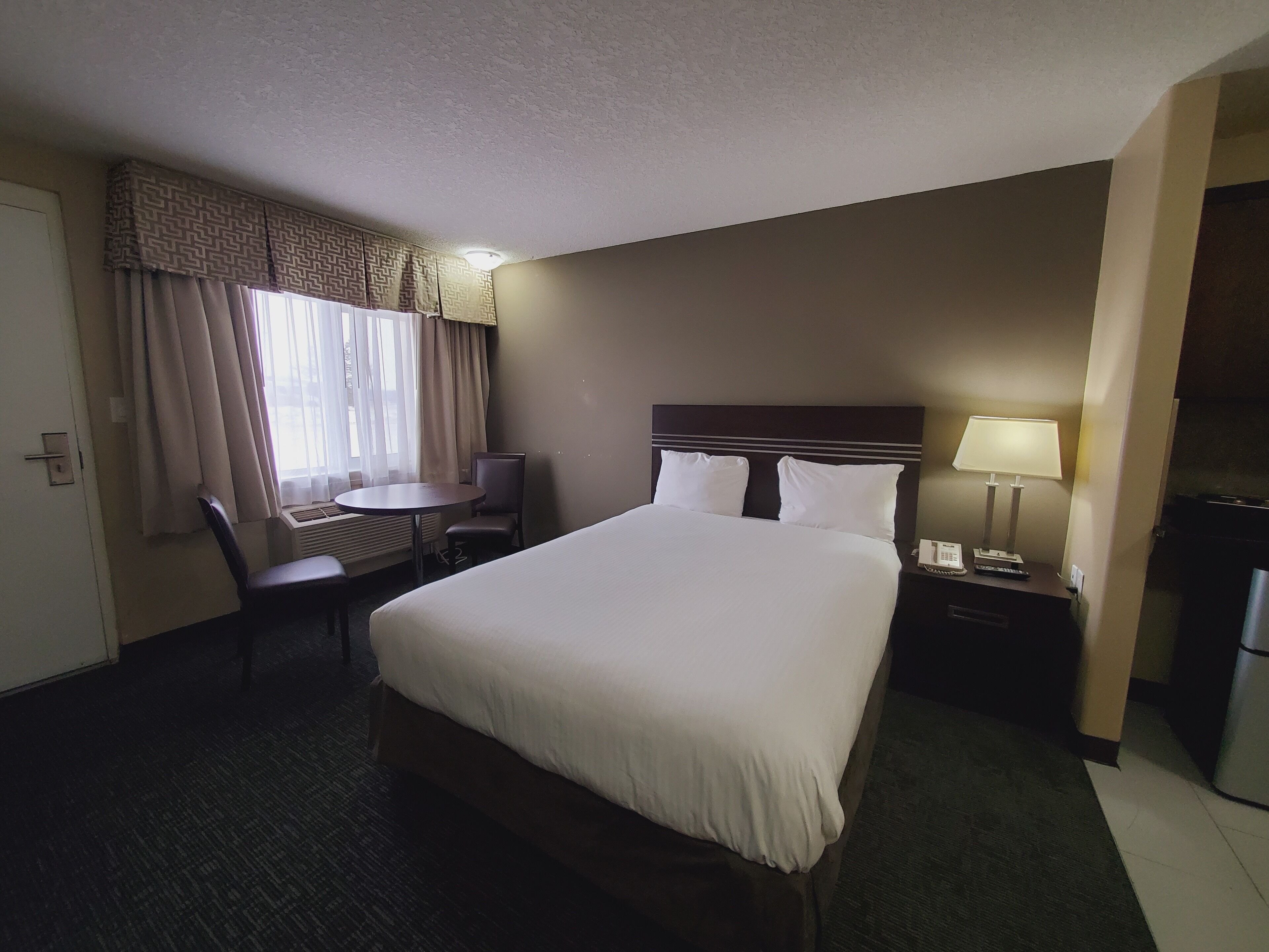A hotel room with a large bed, a desk, a chair, a nightstand with a lamp, and a window with curtains.