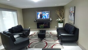 A living room with two black leather sofas, a glass coffee table, a flat-screen TV mounted above a fireplace, and a patterned rug. - Royal Lodge (Edmonton)
