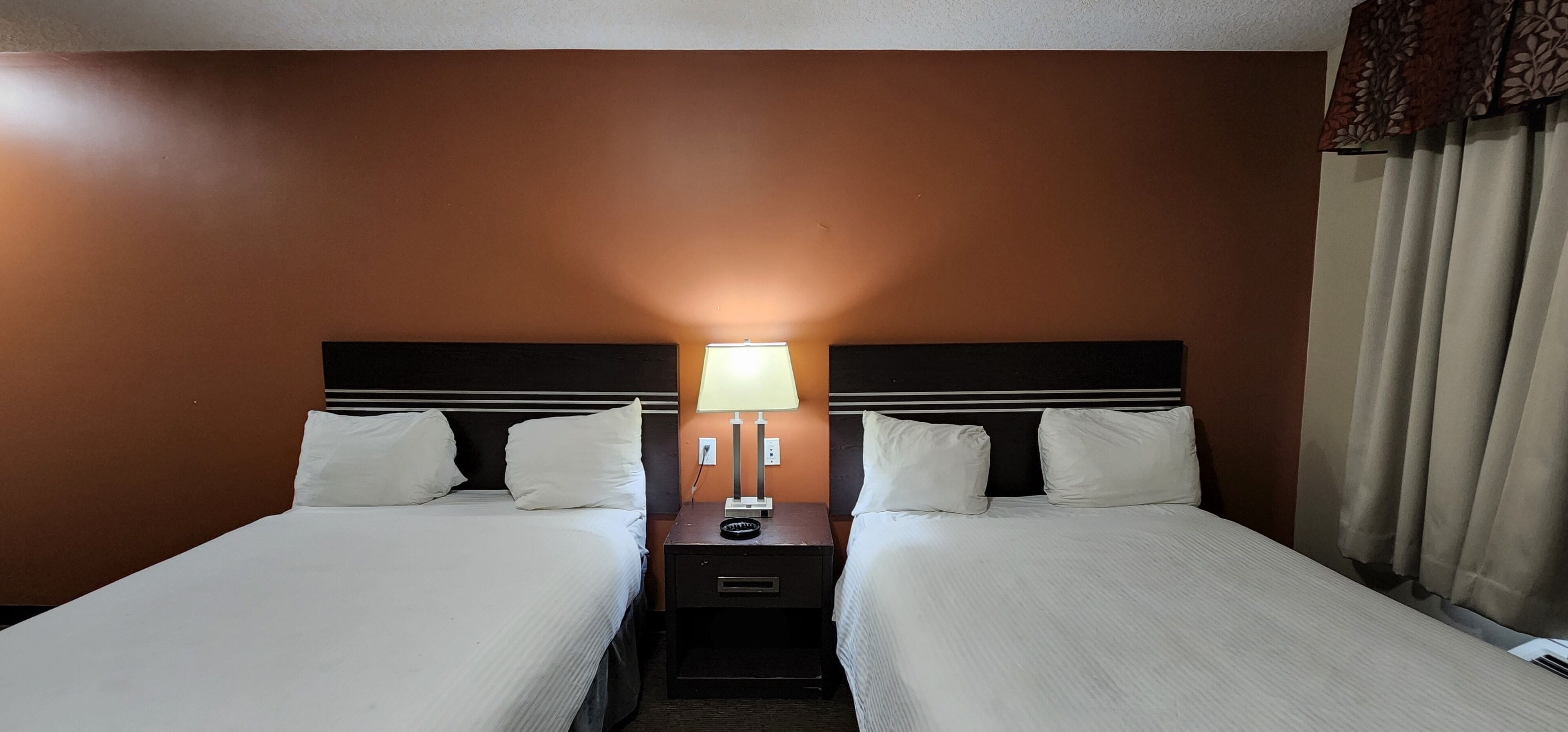 A hotel room with two beds, a nightstand with a lamp, and a window with curtains.