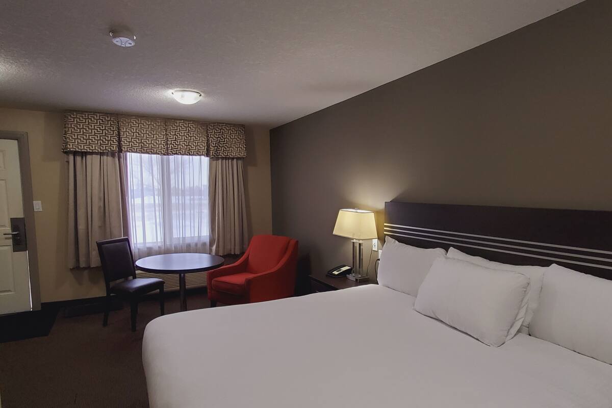 A hotel room with a large bed, a desk with a chair, a red chair, a lamp, and a window with curtains.