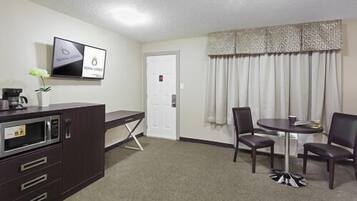 A hotel room with a kitchenette, a dining area with a table and chairs, a TV mounted on the wall, and a door with a window.