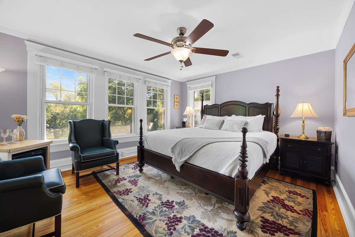 A bedroom with a large bed, a ceiling fan, a nightstand, a chair, and a window with a view of trees.