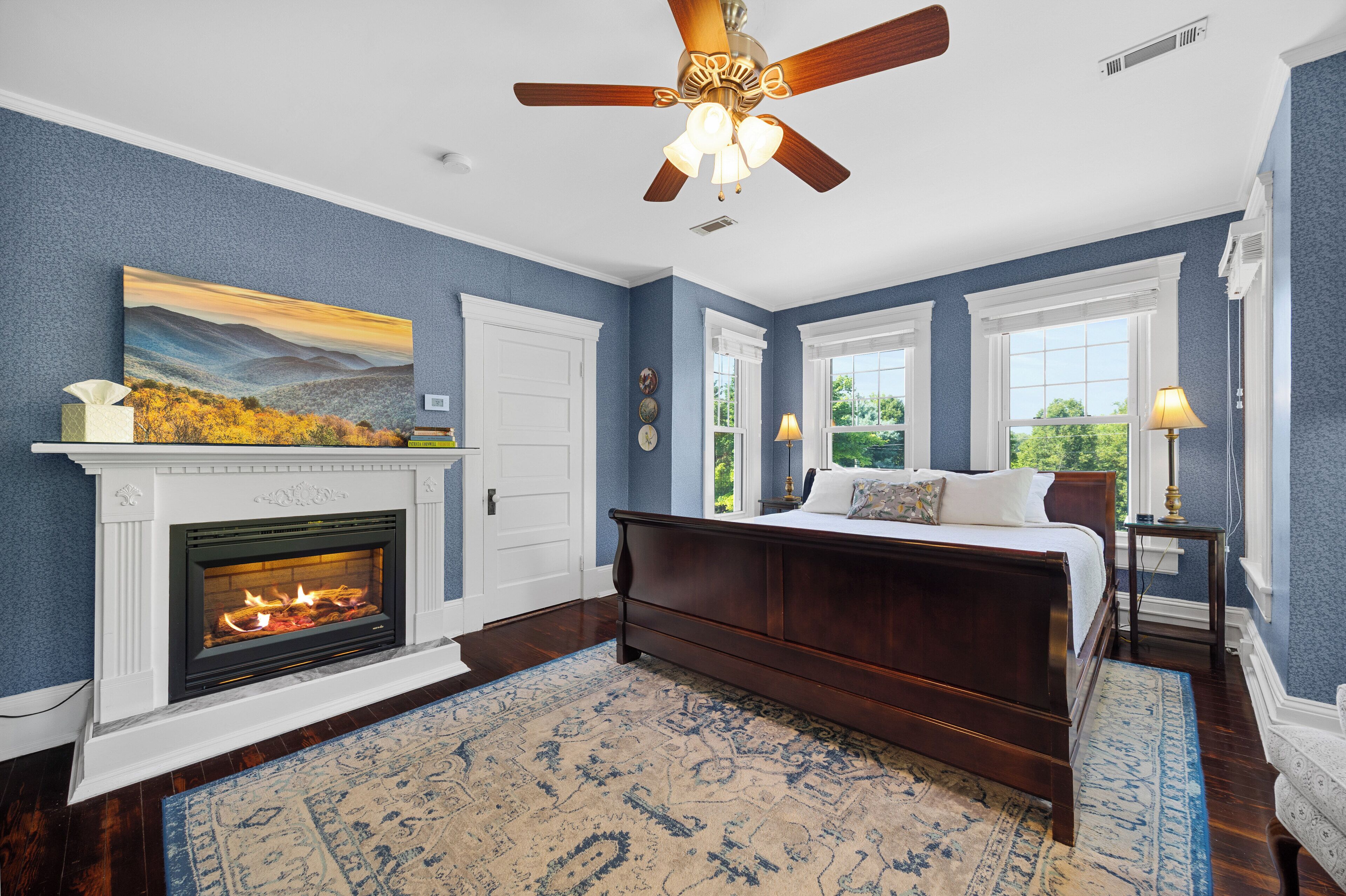 A bedroom with a fireplace, a bed, a ceiling fan, and large windows.