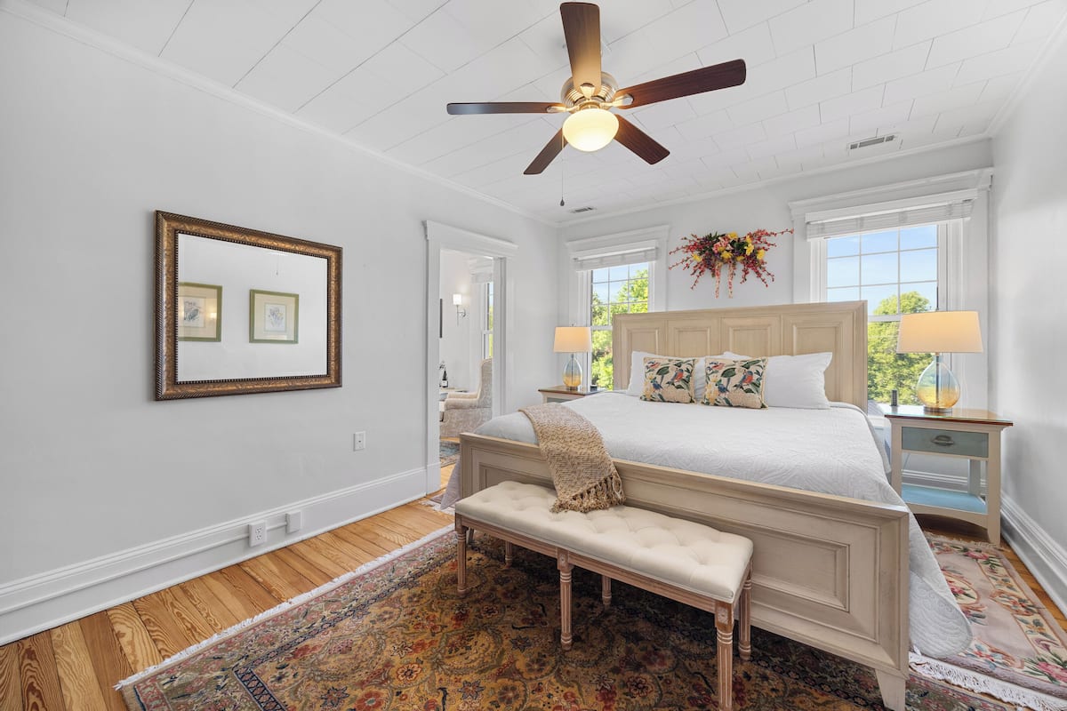 A bedroom with a large bed, a ceiling fan, a bench, and a floral arrangement.