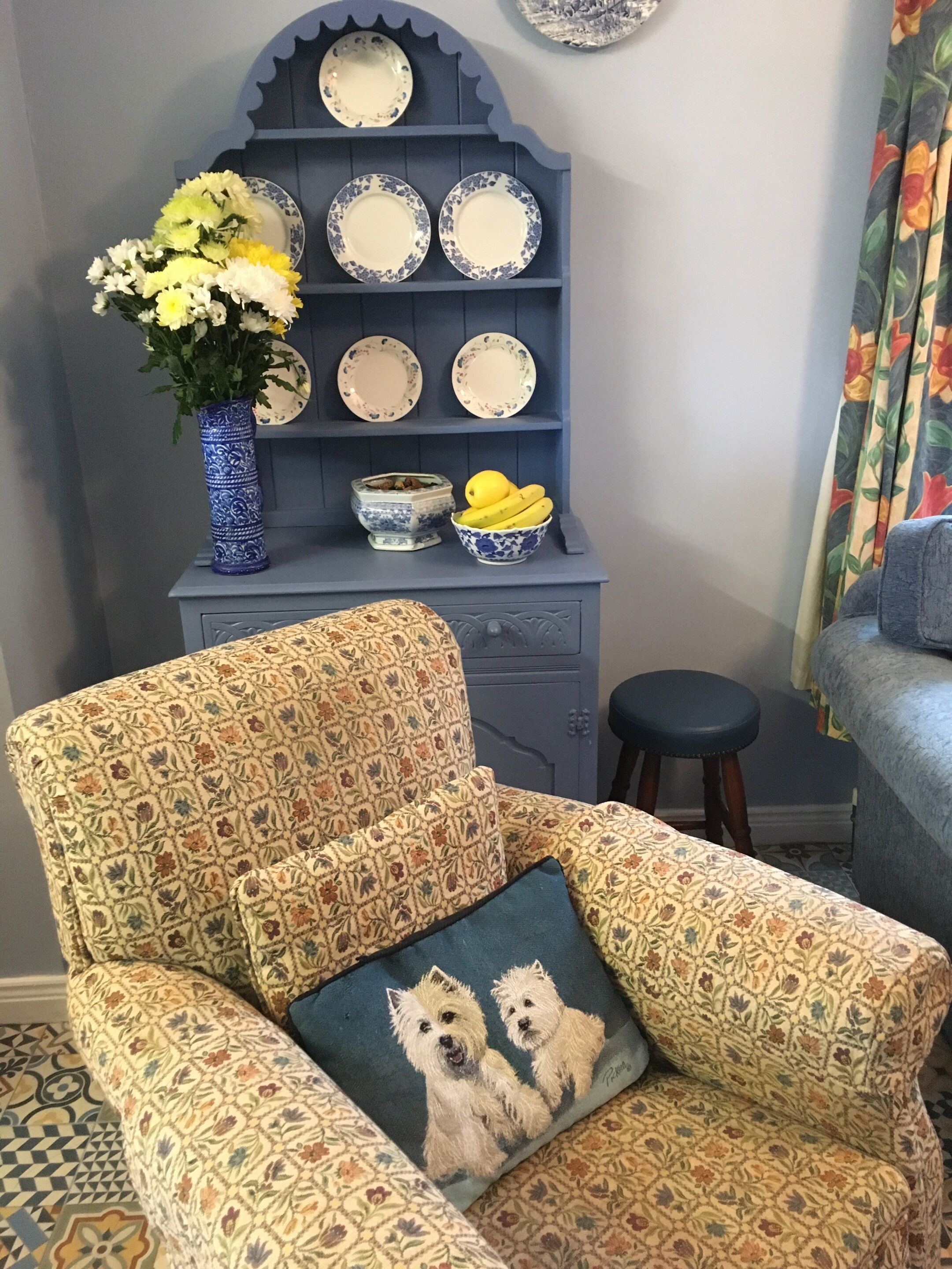 Een gestreept fauteuil met een kussen waarop twee honden afgebeeld zijn, een blauwe vaas met bloemen en een blauwe kast met borden en een schaal met fruit.