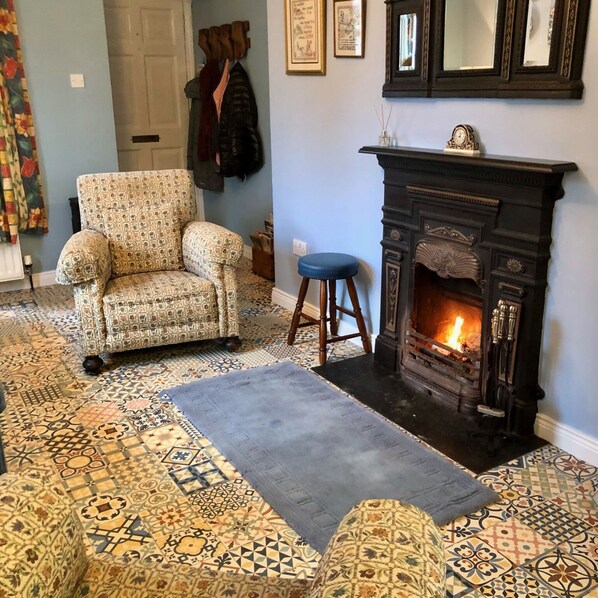 A cozy living room with a patterned carpet, a patterned armchair, a blue stool, and a fireplace.