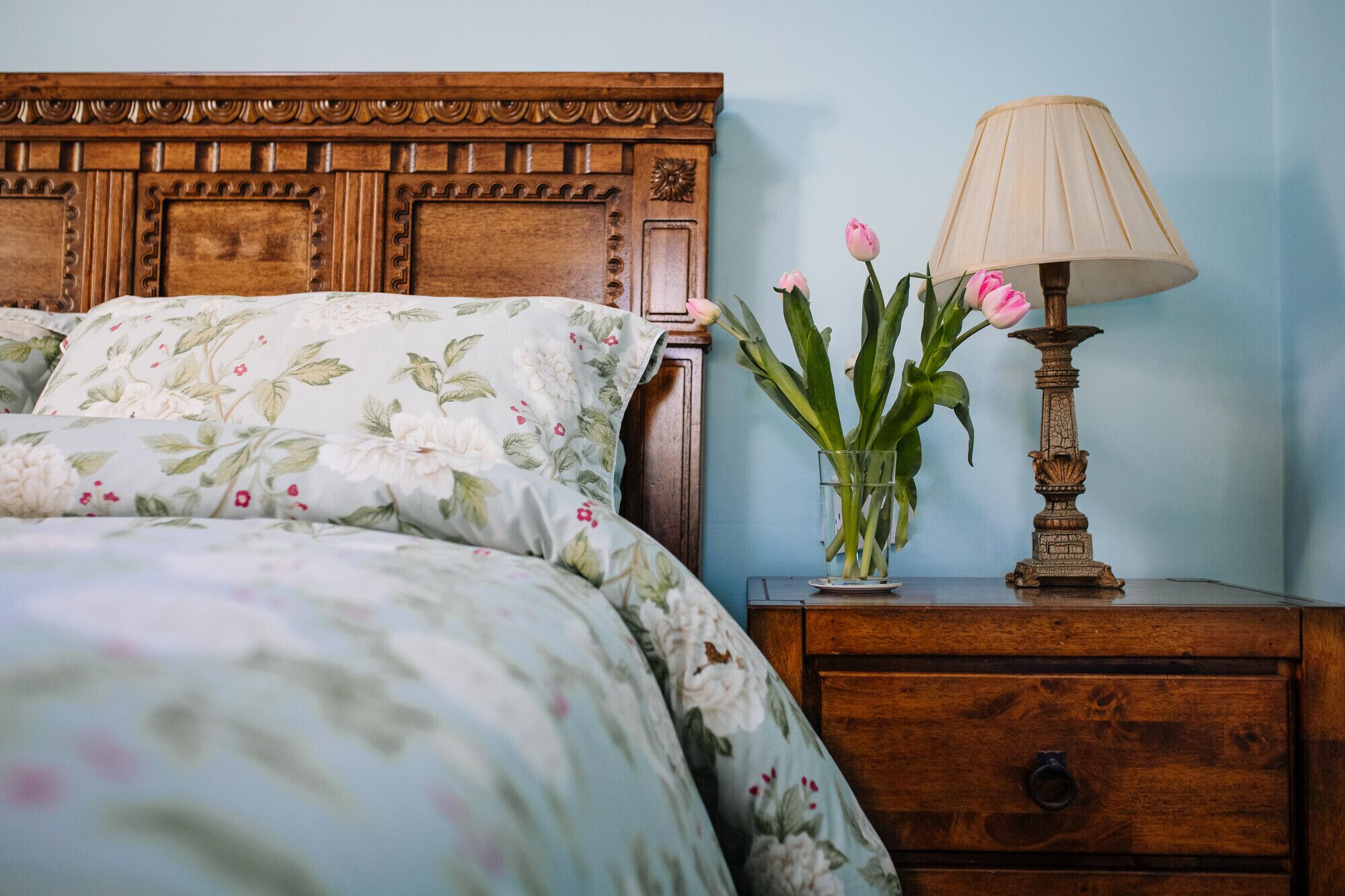 A bedroom with a wooden headboard, floral bedding, a bedside table with a lamp, and a vase with flowers.