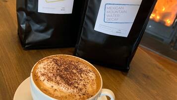 Una taza de café con espuma, dos bolsas de café con las etiquetas 'John Watt Espresso 1865' y 'John Watt Mexican Mountain Water Decaf', y una chimenea al fondo.