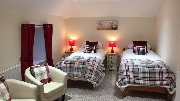 A hotel room with two beds, each with a plaid bedspread, a red lamp on each nightstand, a white chair, and a painting on the wall. - The Village Inn and Kirtle House B&B (Lockerbie)