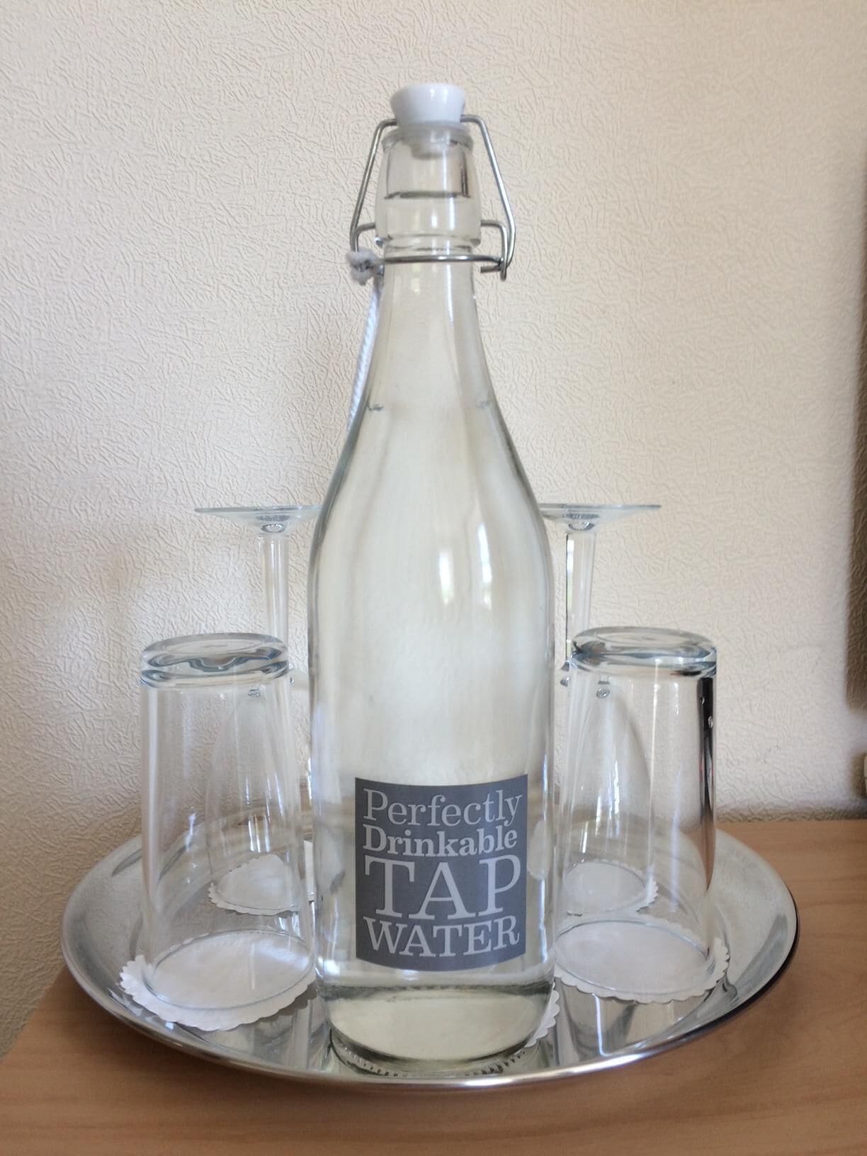Eine Glasflasche mit dem Aufdruck "Perfectly Drinkable TAP WATER“ und drei Gläser auf einer Tablett.