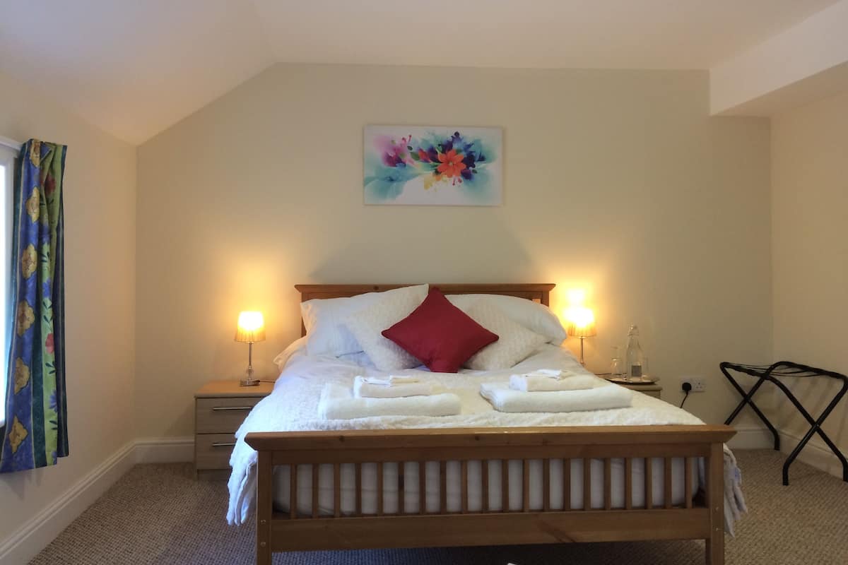 A neatly made bed with white linens and a red pillow, two bedside lamps, a small bedside table, and a colorful abstract painting on the wall.