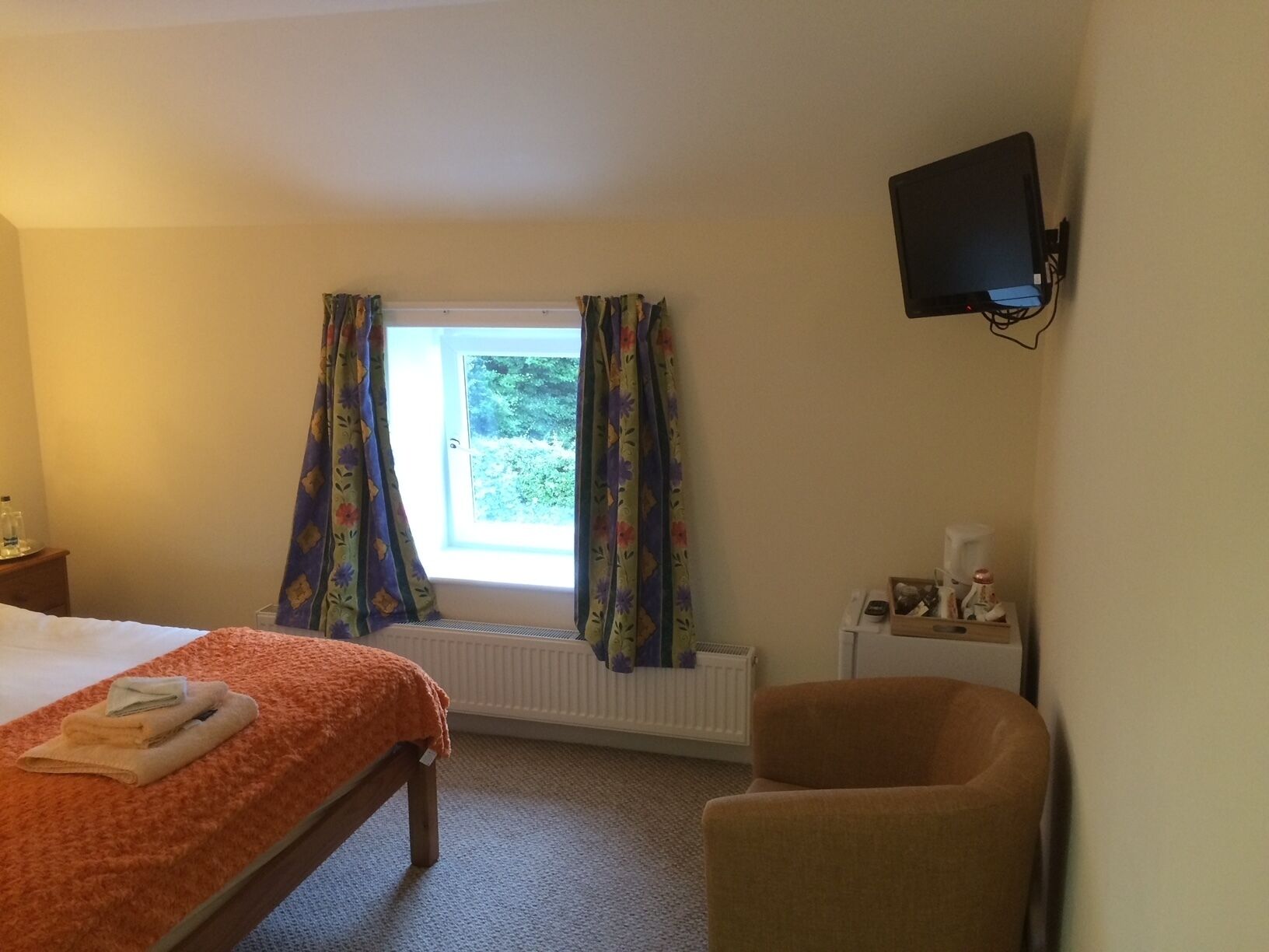 Habitación de hotel con cama, una silla, ventana con cortinas, radiador y televisor montado en la pared.